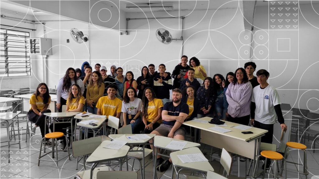 Foto com diversos jovens em uma sala de aula. Alguns usam a camiseta amarela com logo do Semear. Todos olham para a câmera e sorriem. Eles estão em uma sala de aula com mesas e cadeiras beges que estão espalhadas ao seu redor.