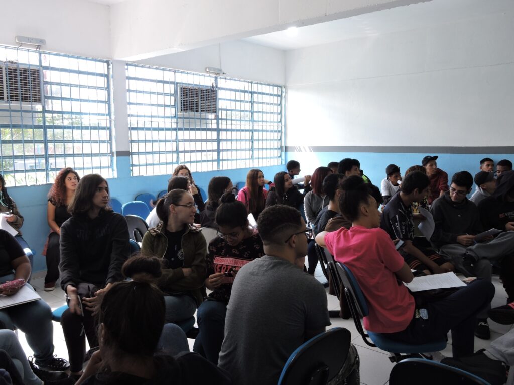 A foto mostra diversos jovens em uma sala de aula azul, todos estão sentados em cadeiras beges e olhando para o professor que não aparece na foto.