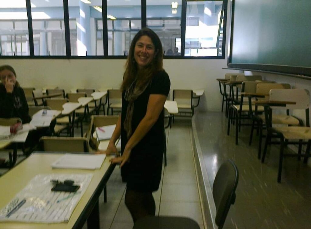 Em foco na foto está a professora Camila Medina, uma mulher branca de cabelos castanhos na altura dos ombros sorrindo para a câmera. Ela usa camiseta e calça pretas e está em frente a mesa da professora. A foto mostra ao fundo uma sala de aula vazia, com mesas e cadeiras beges e uma janela grande ao fundo.